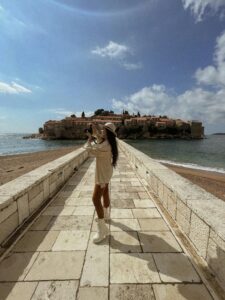 A woman photographs the scenic Sveti Stefan connected by a paved walkway in Montenegro.
