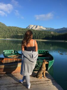 A woman and her dog stand on a dock overlooking a tranquil mountain lake.